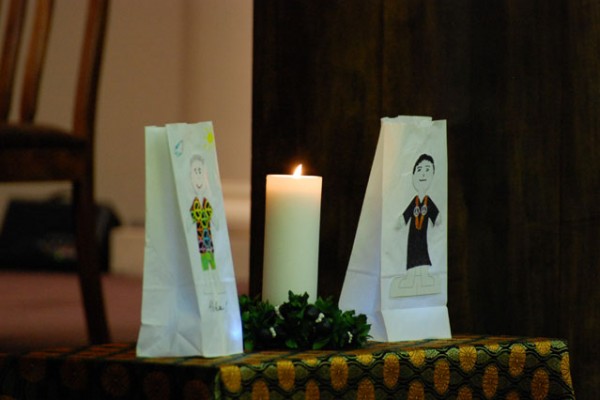 white paper sacks on either side of a candle; one with drawing of man in Aloha shirt with peace symbols, one with drawing of minister