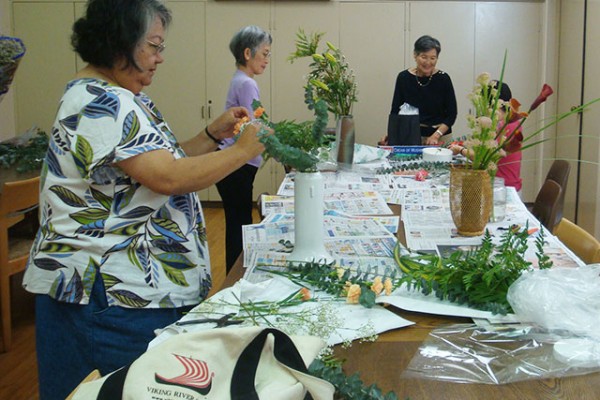 Women standing at a table in the social hall arrange flowers with the instructor Edith Tanaka looking on.