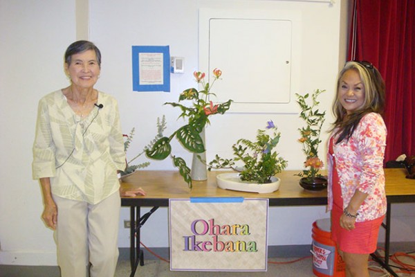two women at a table with flower arrangements and a sign reading "Ohara Ikebana"