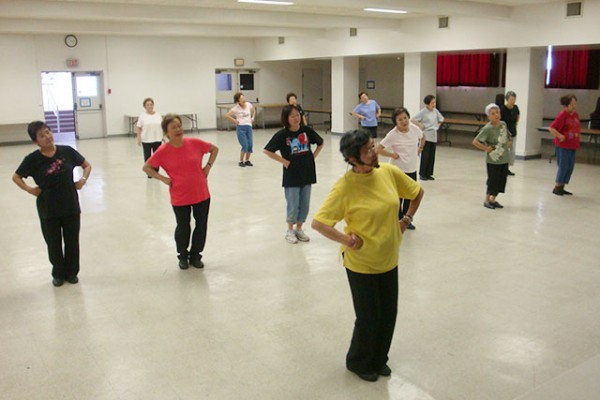adult students spread out in the social hall all face forward to mirror the moves of the instructor