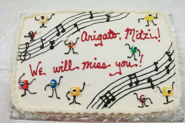 top view of a cake decorated with musical bars and notes and the words, "Arigato, Mitzi! We will miss you!"