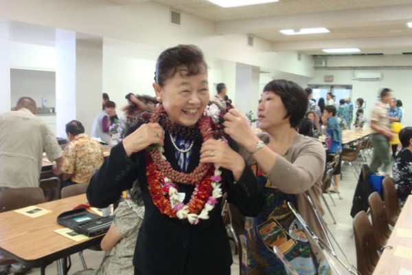 Mrs. Saito smiling and wearing leis assisted by a young woman