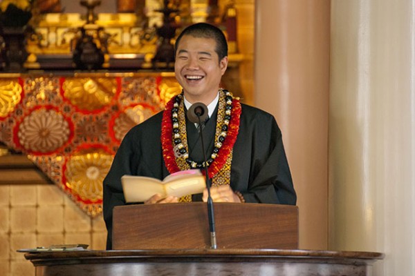 Rev. Jay Okamoto at the pulpit with leis and a big smile holding a chidren's book