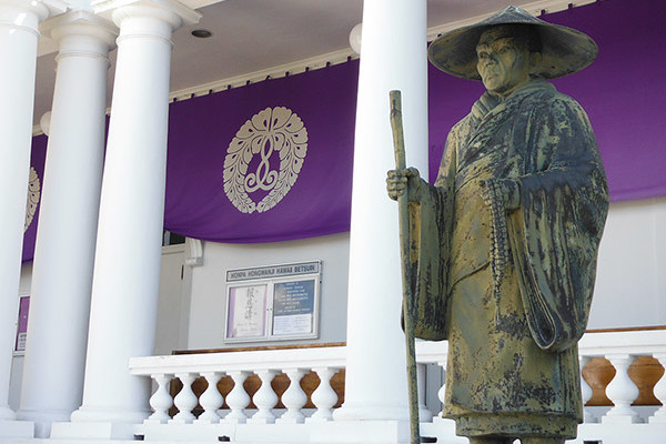 Shinran Shonin statue with Hawaii Betsuin hondo entrance and purple major service banner with wisteria logo in background