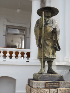 Shinran Shonin statue with plaque; white building and corridor of Hawaii Betsuin in the background
