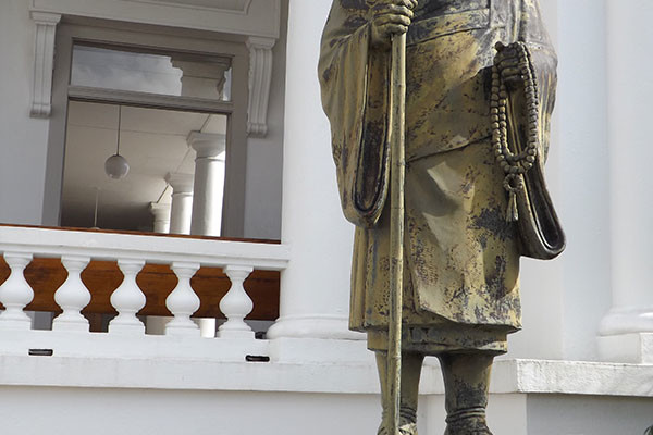 Shinran Shonin statue with plaque; white building and corridor of Hawaii Betsuin in the background