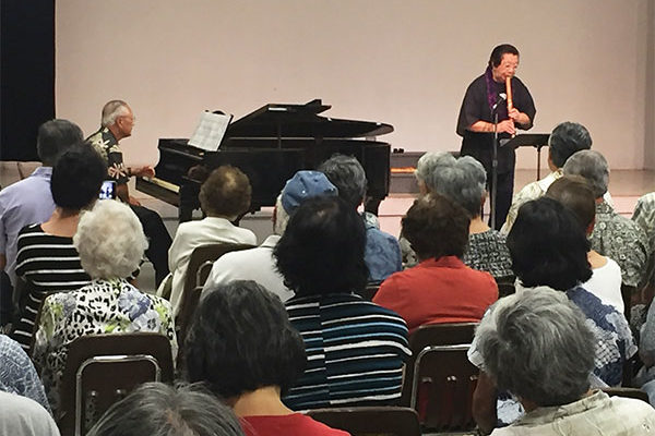 audience watches and listens as Miyoshi Genzan plays the shakuhachi flute and Francis Okano plays piano in the Hawaii Betsuin social hall