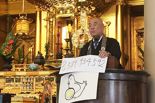 Rimban Hagio at the pulpit with a poster of a chicken drawing