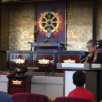 Rev. Jan Youth speaks from the podium in the Annex Temple with the Wheel of Dharma and altar in the background