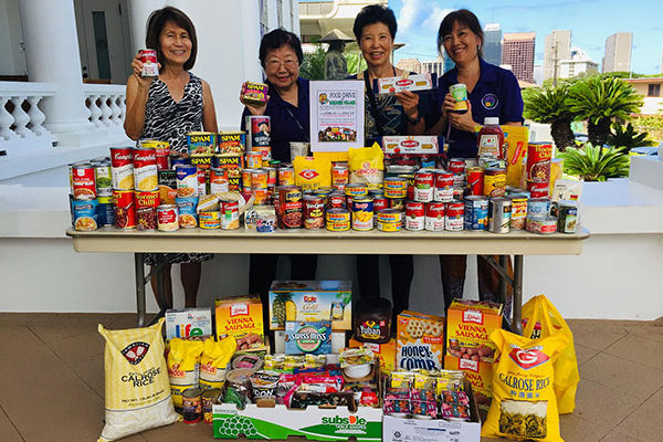 four women stand with stacked cans of food, bags of rice, and other nonperishables outside the Hawaii Betsuin hondo on the last day of the food drive
