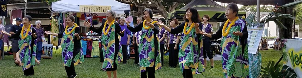 video capture - Hawaii Betsuin choir performs Golden Chain with hula and sign language at VegFest Oahu 2019; women with lei and hands outstretched on grass in front of the stage
